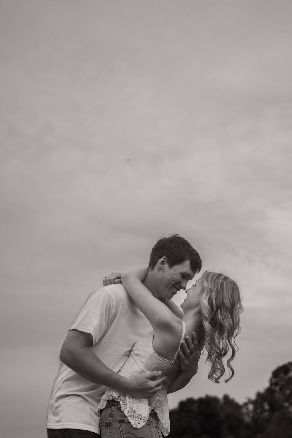 Black and white romantic photo of couple leaning in for a kiss against a cloudy sky backdrop, engagement photographer Greensboro NC