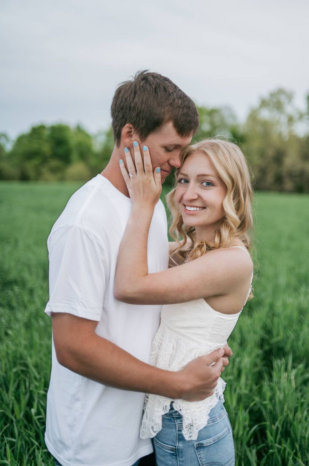 Smiling blonde woman embraces her partner in a lush green field during an outdoor engagement session, High Point NC
