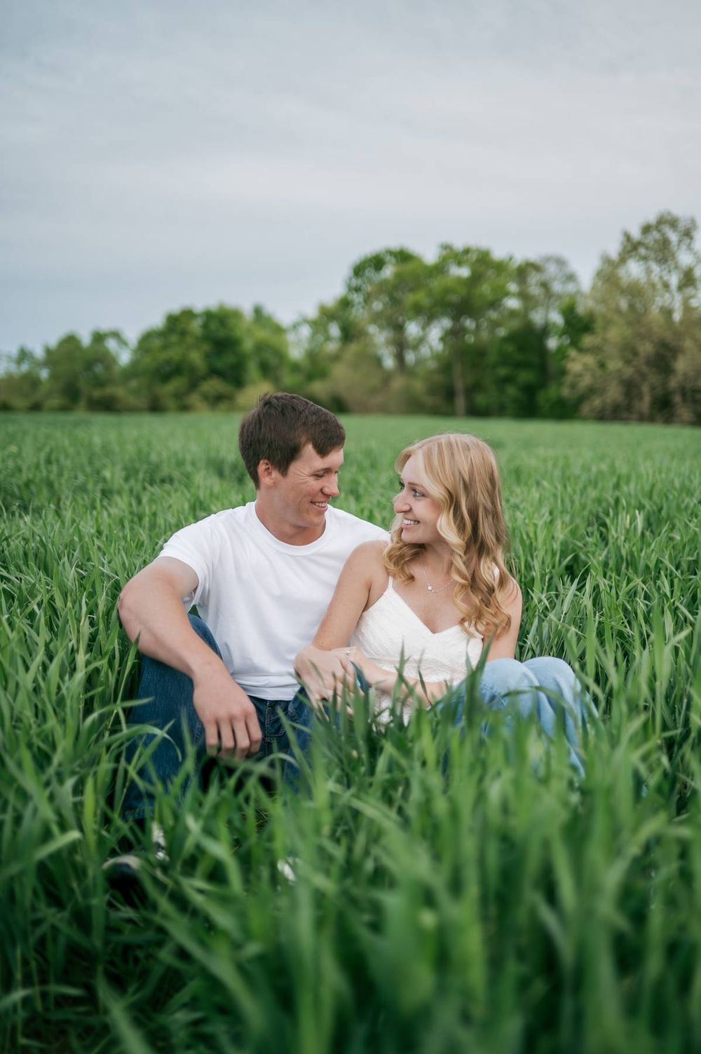 Happy couple sitting together in tall green grass, smiling at each other during engagement photo session, Asheboro NC