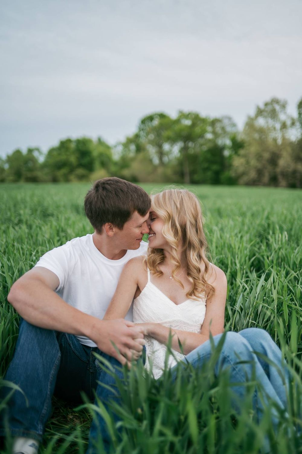 Romantic couple sitting in a green field leaning in for a kiss during their engagement photography session, engagement photographer NC