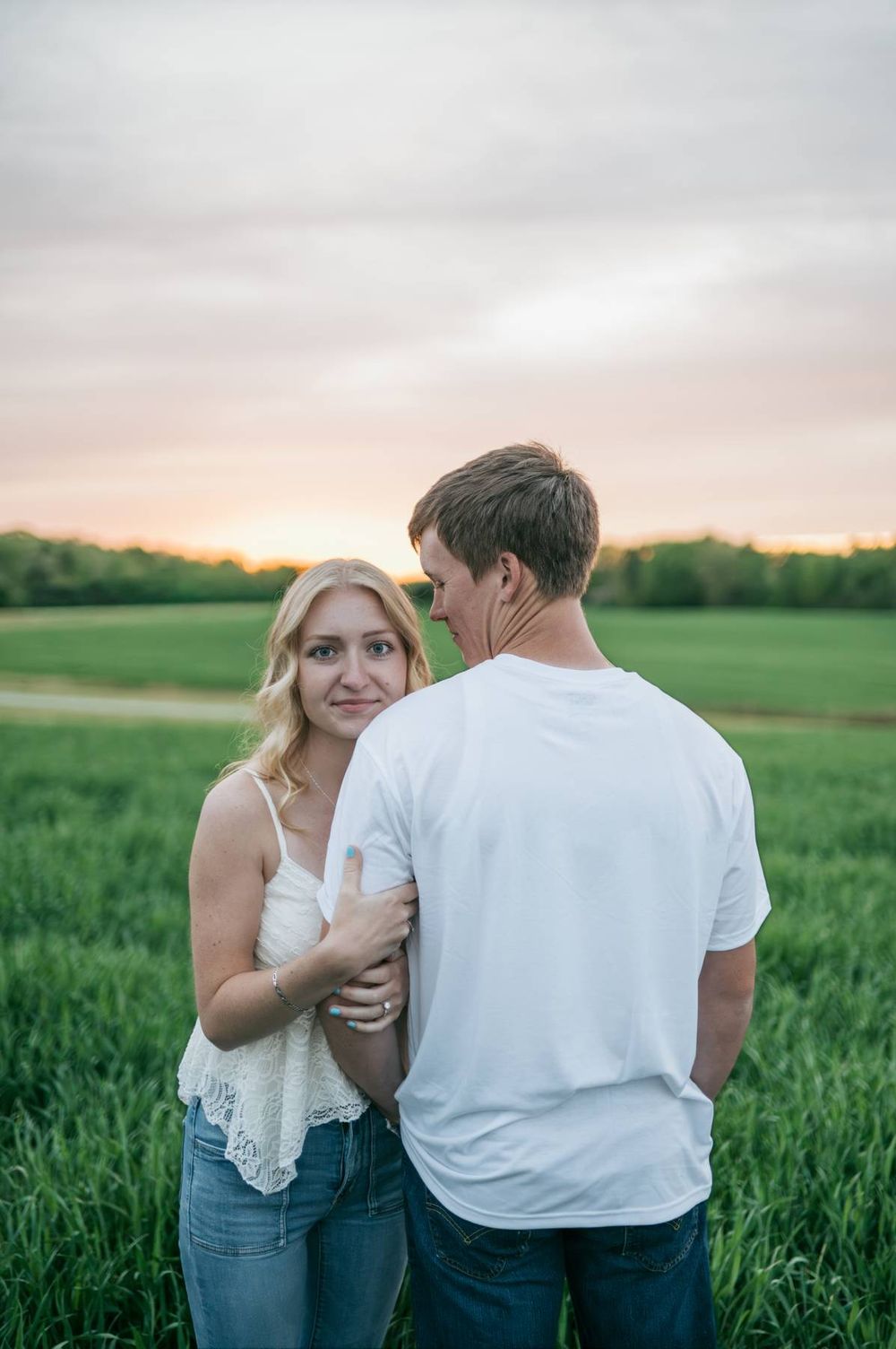 Blonde woman smiles at camera while embracing her partner in a green field at sunset, engagement photographer Greensboro NC