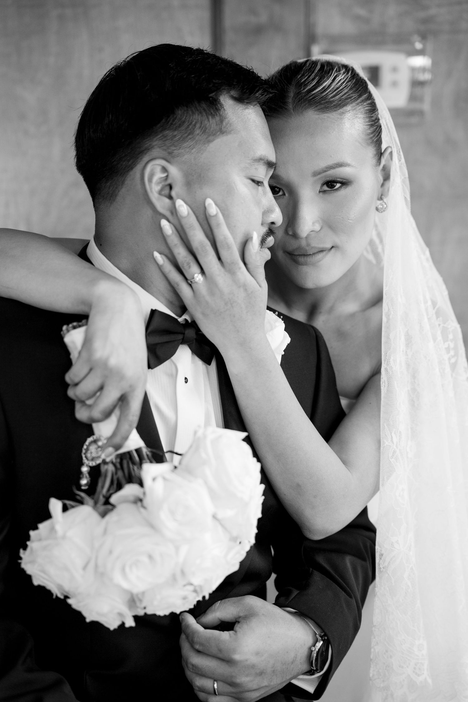 Black and white portrait of bride tenderly holding grooms face while embracing with a white rose bouquet.