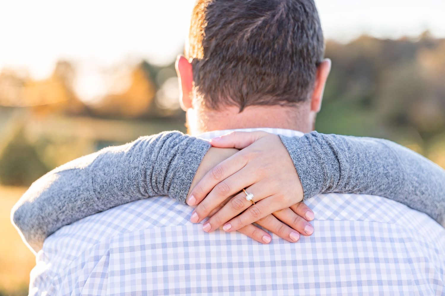 Couple embracing outdoors at sunset, woman wearing diamond engagement ring on man's shoulder.