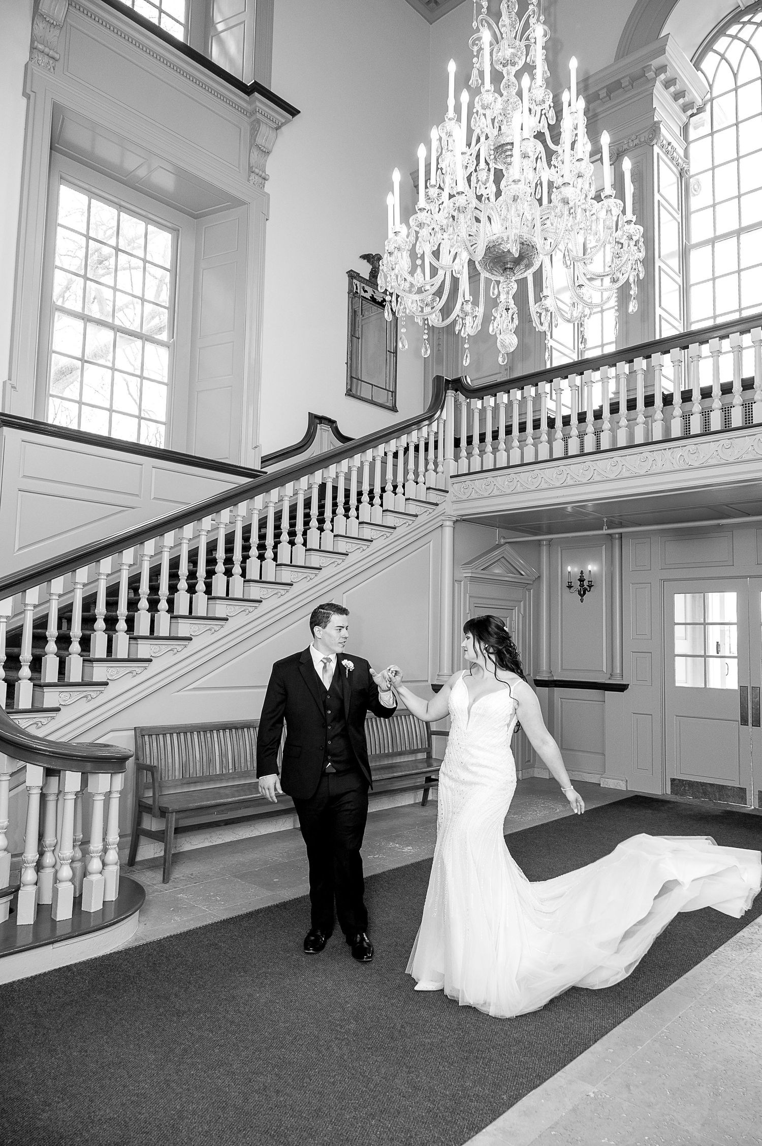 Black and white photo of bride and groom dancing in elegant venue with grand staircase and crystal chandelier.