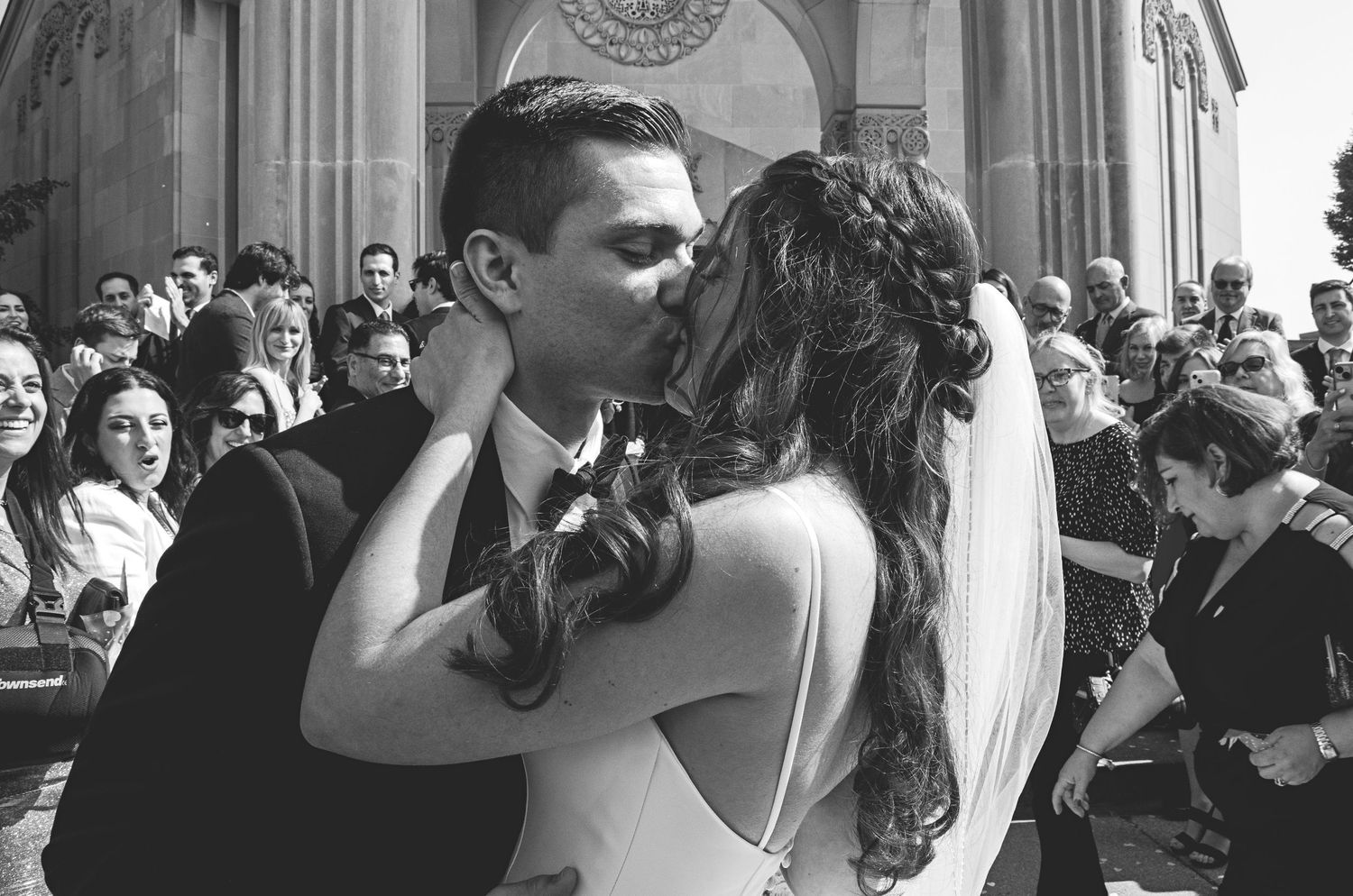 Black and white photo of a bride and groom sharing their first kiss outside a church surrounded by wedding guests.