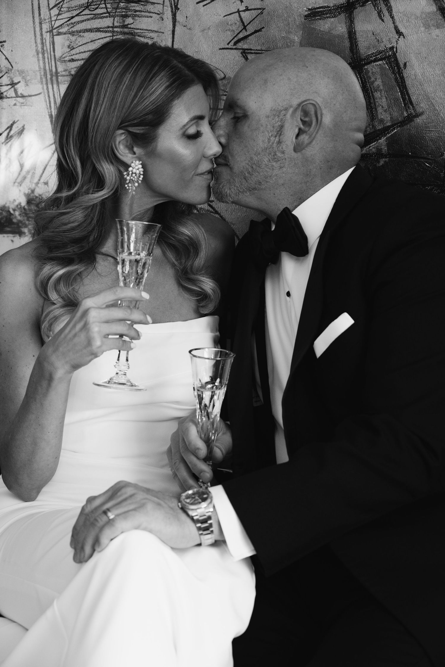 Black and white photo of a couple in wedding attire sharing a kiss while holding champagne glasses.