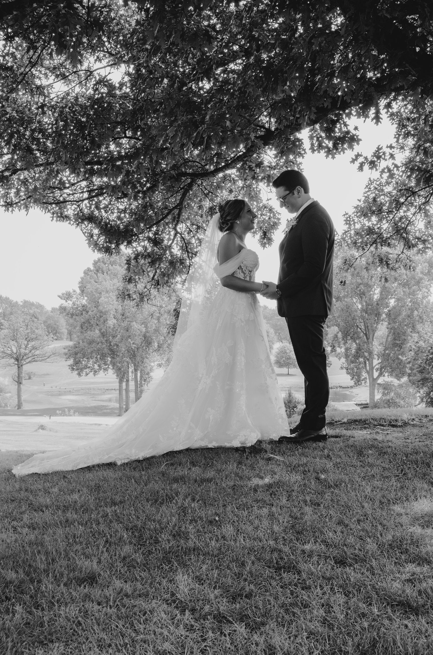 Black and white photo of a bride and groom holding hands under a large tree on their wedding day.