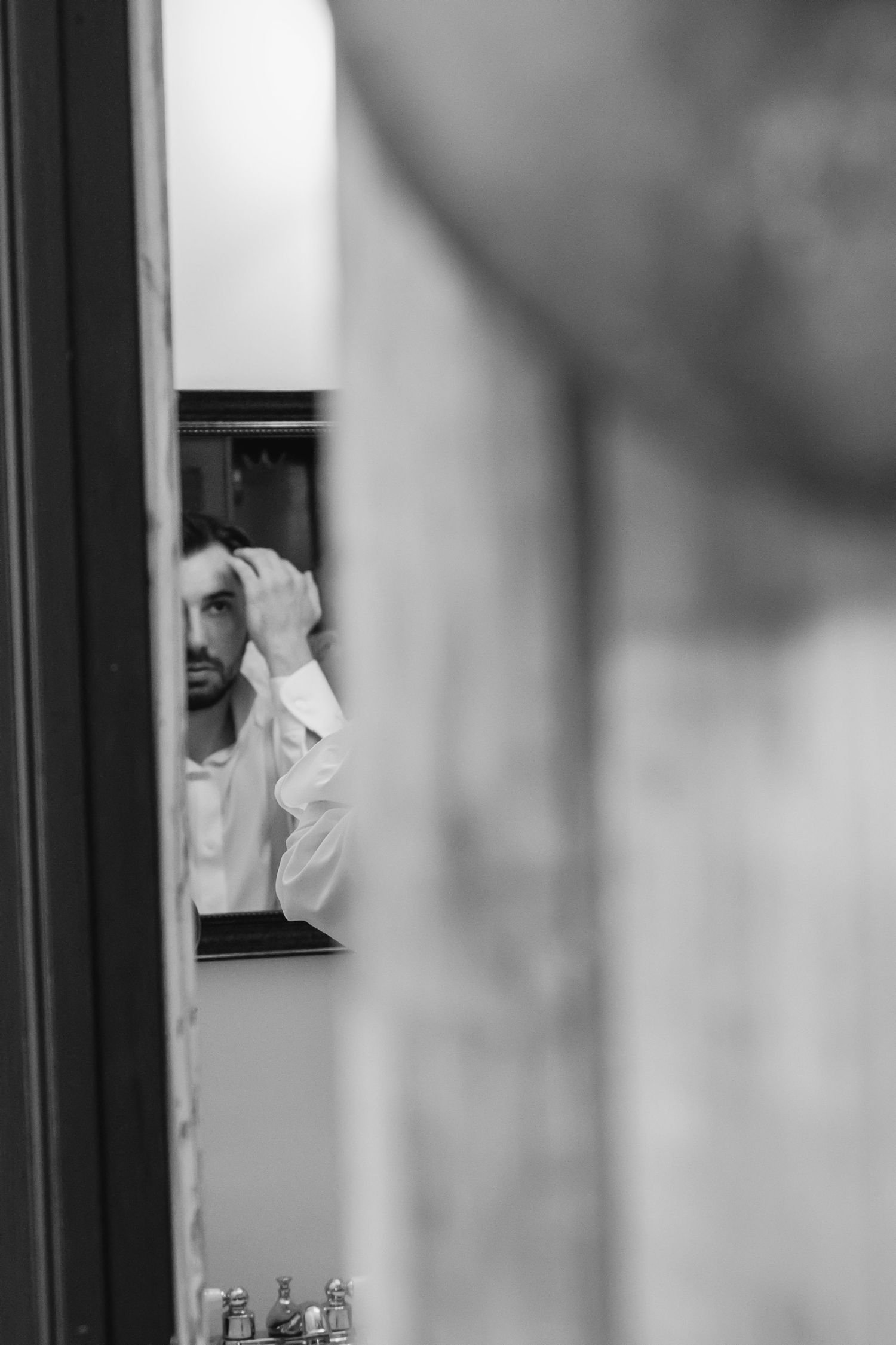 Black and white photo of a man in a white shirt fixing his hair while looking in a bathroom mirror.