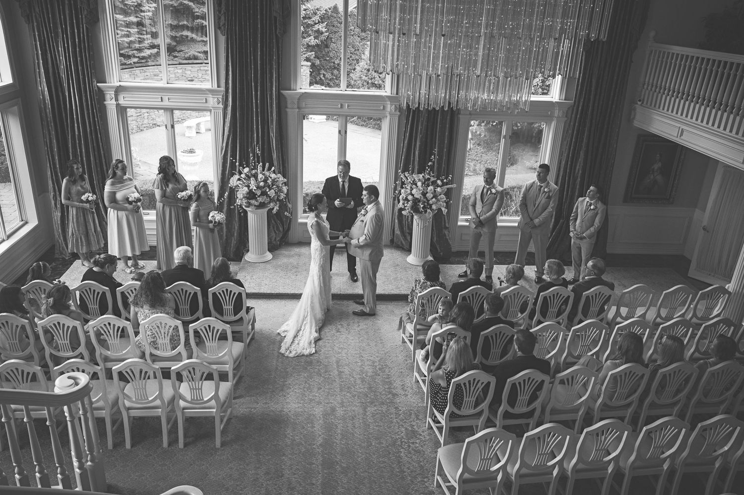 Black and white overhead view of an elegant indoor wedding ceremony with guests seated in ornate chairs.