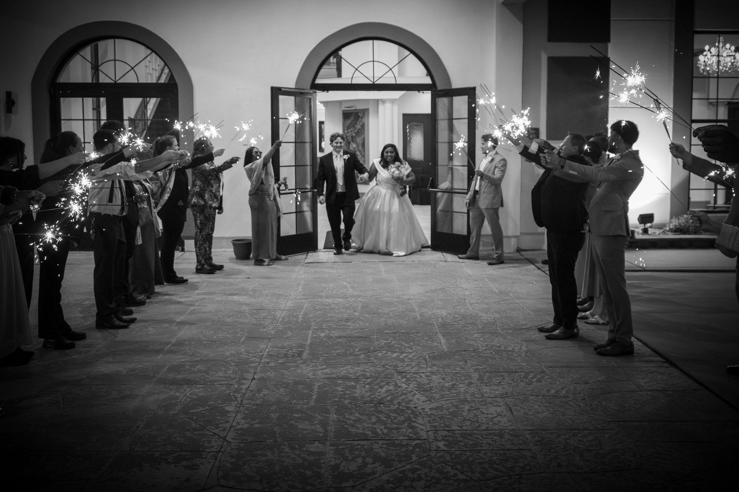 Black and white photo of newlyweds exiting through sparkler send-off tunnel held by wedding guests at night.
