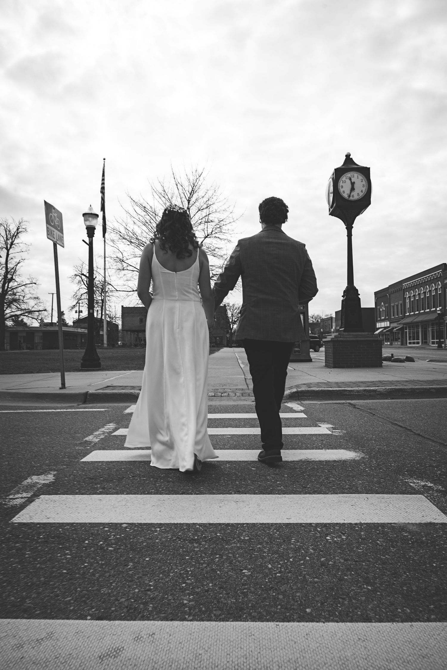 Black and white photo of a bride and groom holding hands while crossing a zebra crossing near a street clock.