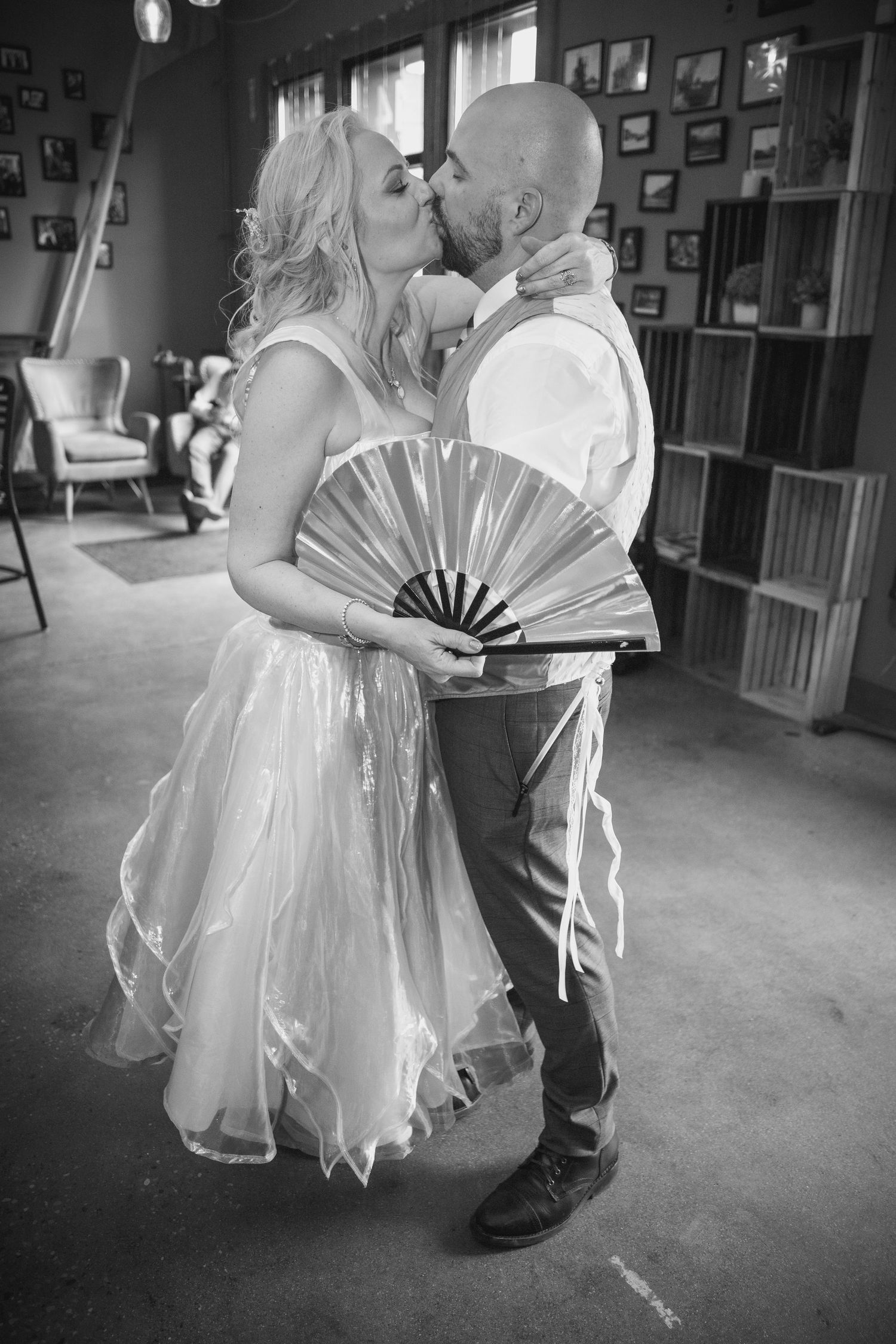 Black and white photo of a bride and groom kissing, bride holding a decorative fan, in a rustic indoor venue.