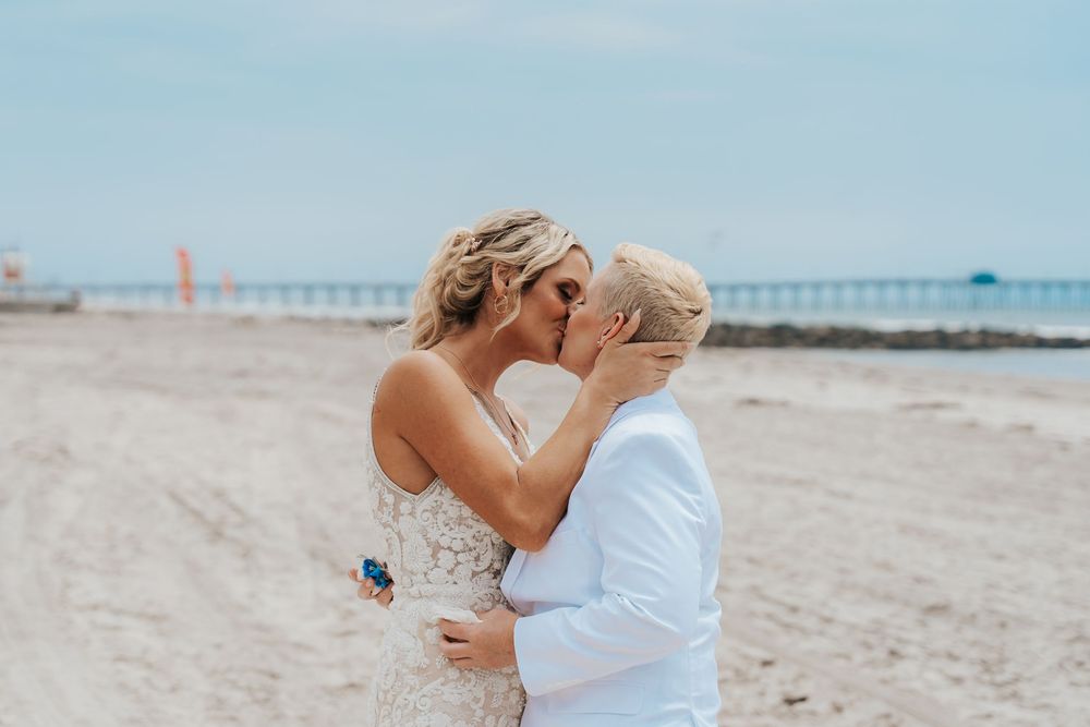 Two brides share a romantic kiss on a sandy beach during their wedding ceremony with a pier in the background.