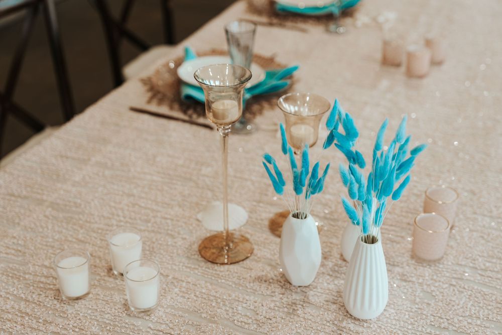 Elegant table setting with gold candelabra, white vases with blue dried flowers, and glowing votive candles on a sequined tablecloth.