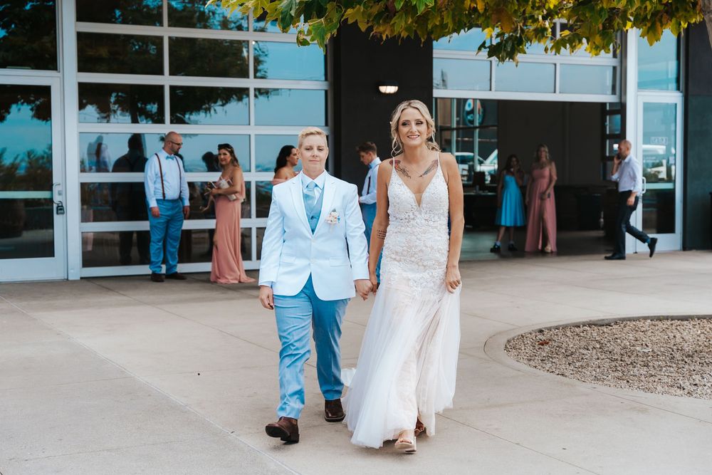 Two brides walking hand in hand outside a modern venue, one in a white gown and one in a light blue suit.
