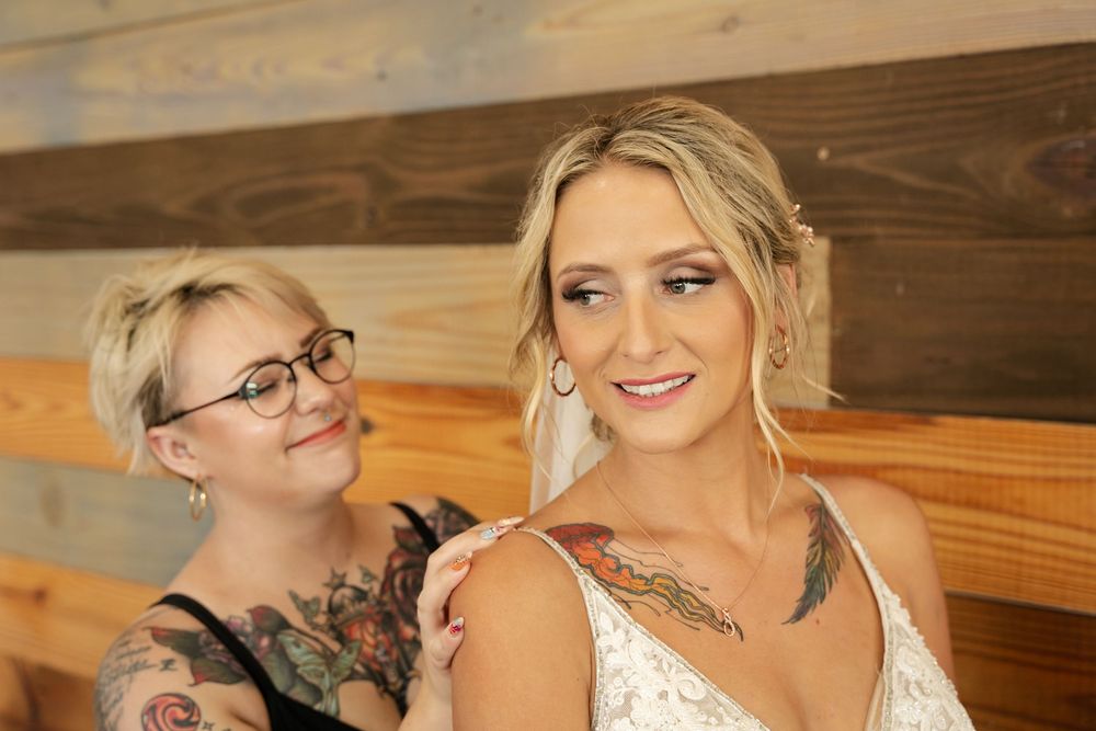 Bride in white lace gown gets final touches from tattooed stylist in rustic wood-paneled venue.