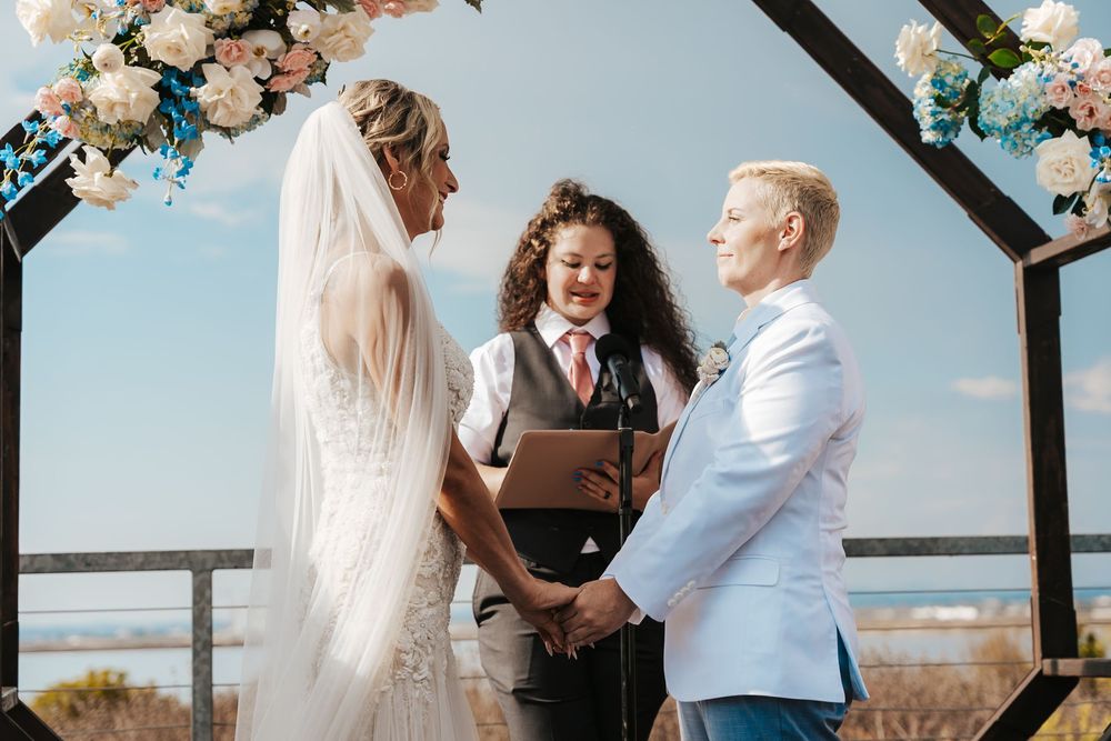 Two brides hold hands during an outdoor wedding ceremony officiated by a smiling officiant under a floral arch.
