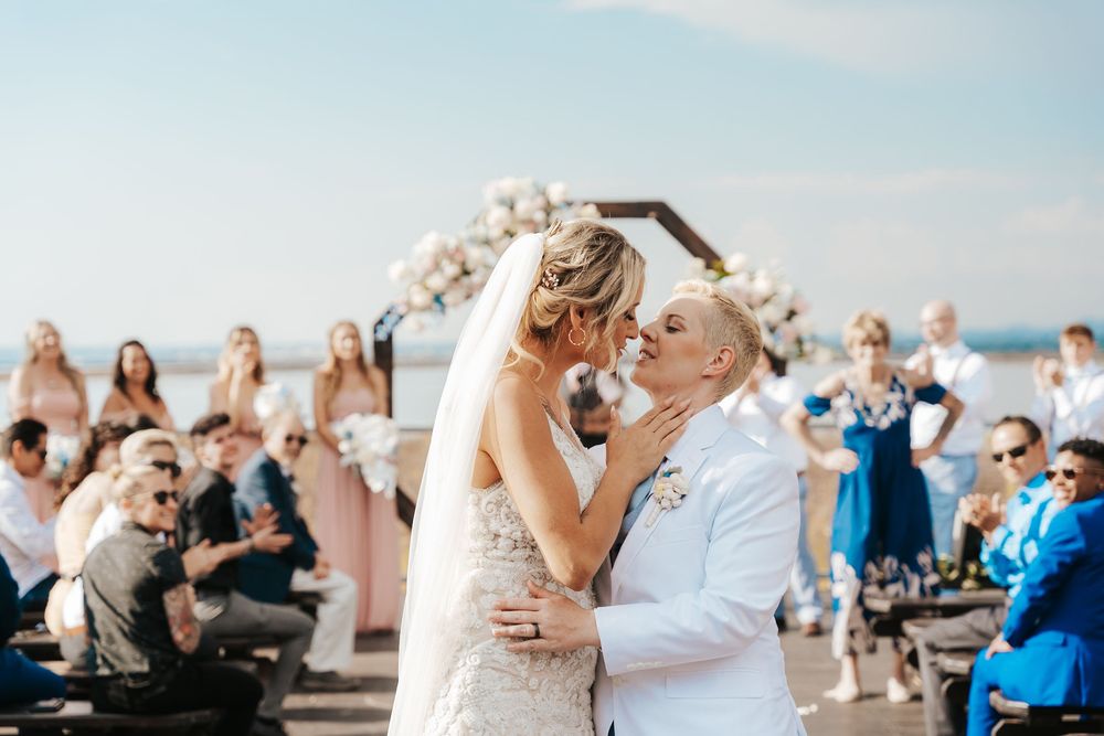 Two brides share their first kiss at an outdoor waterfront wedding ceremony with bridesmaids and guests in the background.