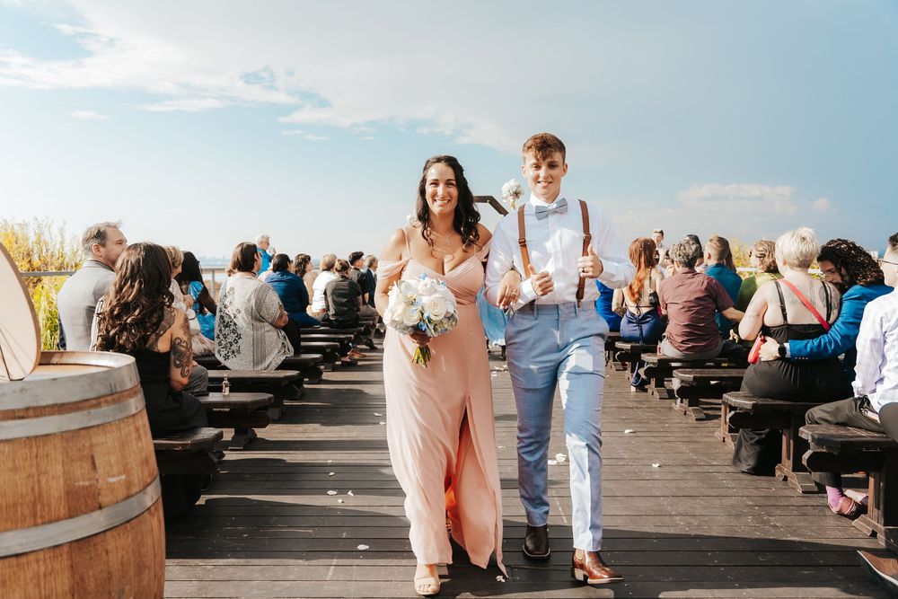 Bride and groomsman walk down outdoor wedding aisle on wooden dock, guests seated on benches under blue sky.