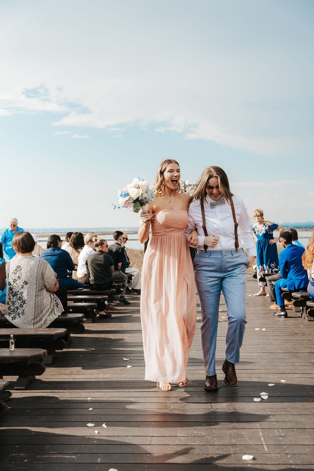 Bridesmaid in pink gown and groomsman in suspenders walk down outdoor wedding aisle on wooden boardwalk.