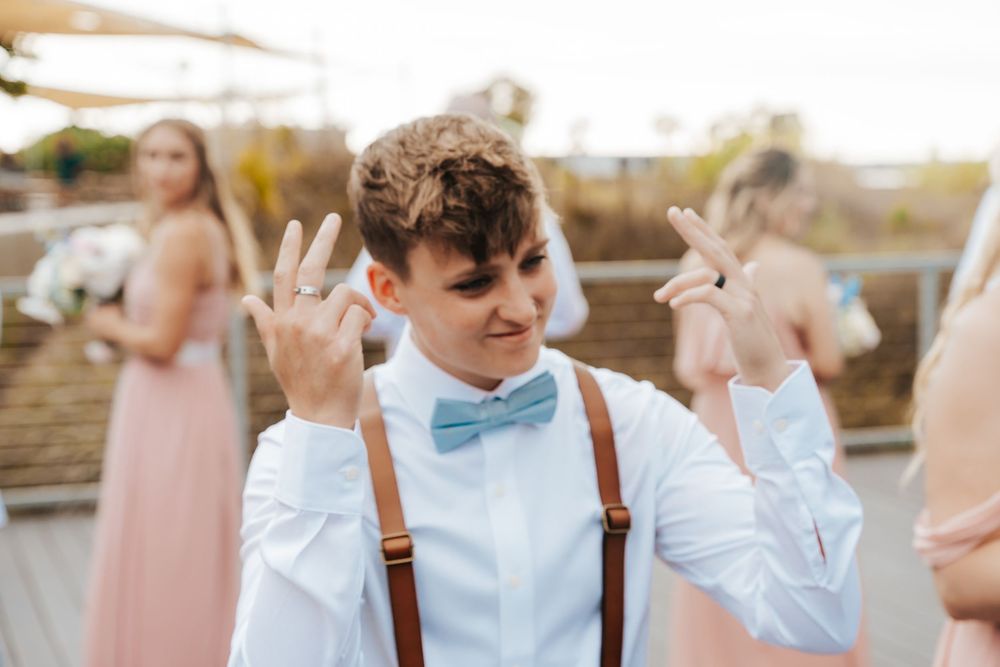 Young man in white shirt, blue bow tie, and suspenders making hand gestures at a wedding celebration.