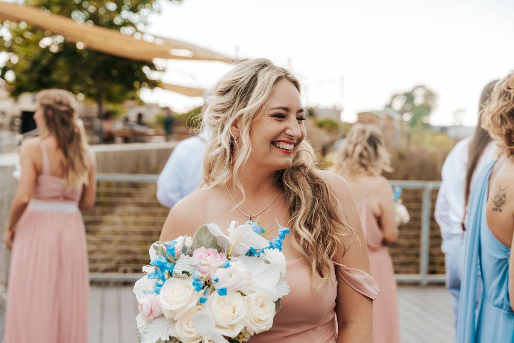 Smiling bridesmaid in pink off-shoulder dress holding white and blue bouquet at outdoor wedding celebration.