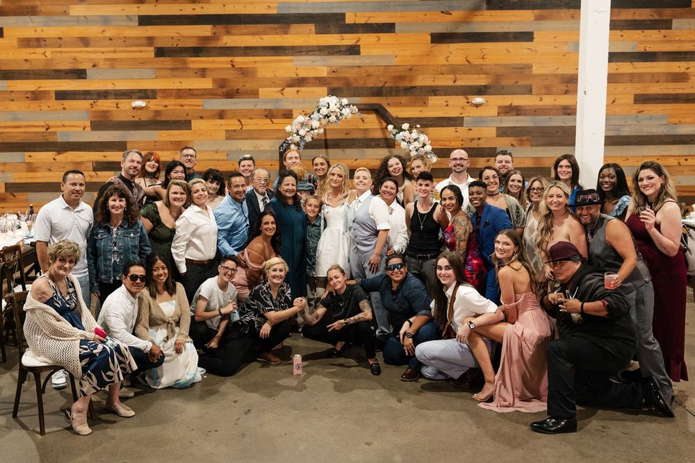 Large wedding group photo with guests posing together in front of a floral arch in a rustic venue.