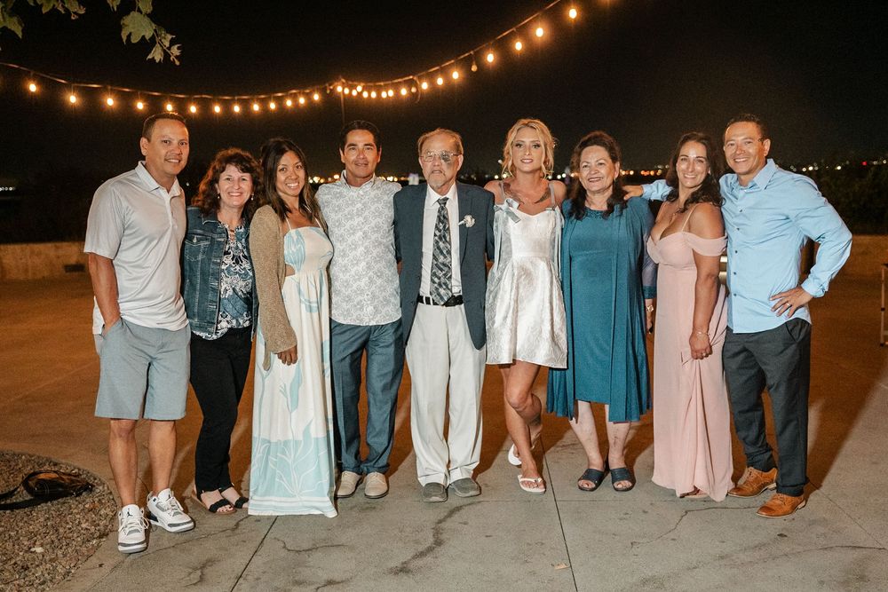 Group of nine people smiling together outdoors at a nighttime wedding reception under string lights.