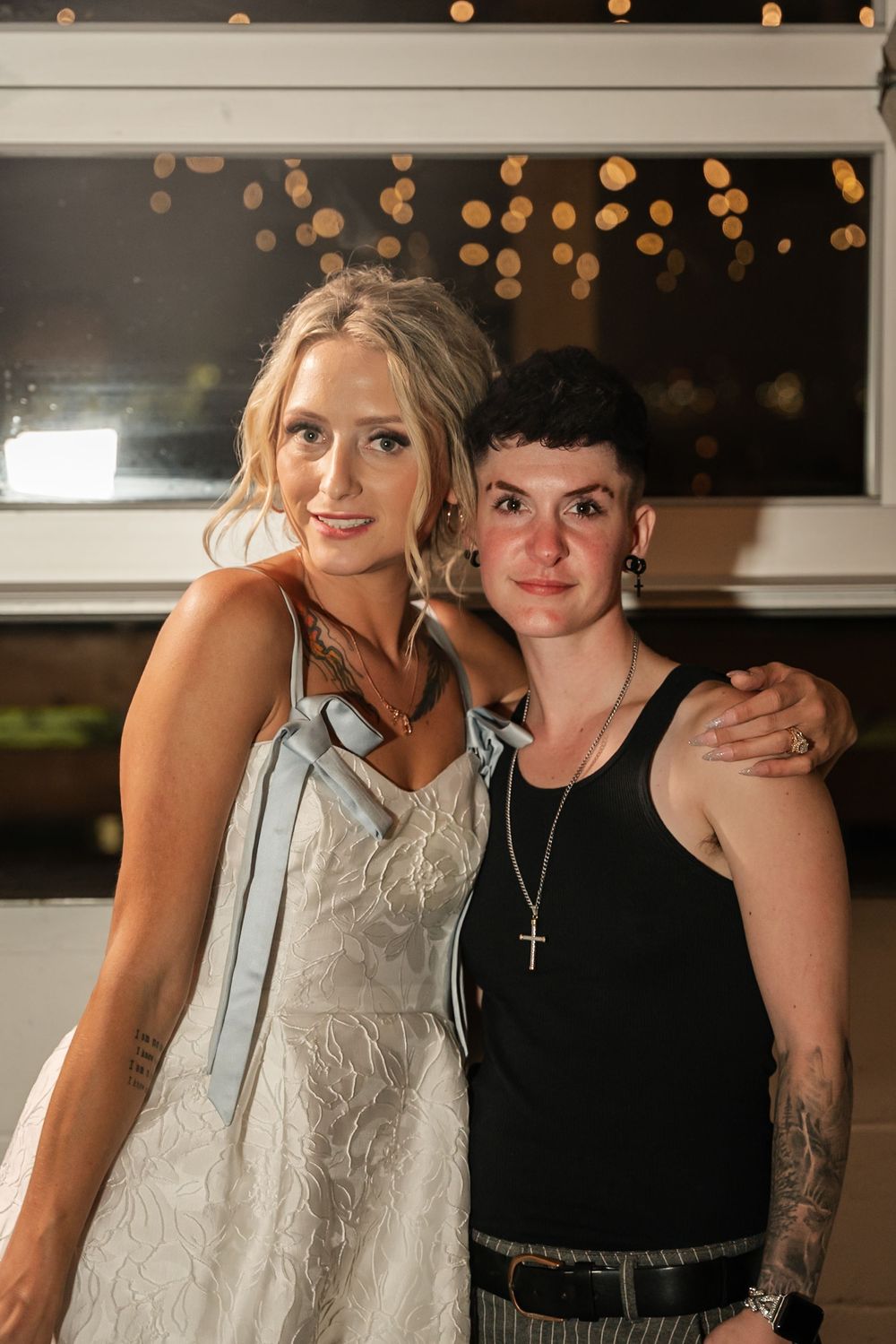 Two women posing together at a wedding reception, one in a white bridal gown with blue ribbon, bokeh lights behind them.