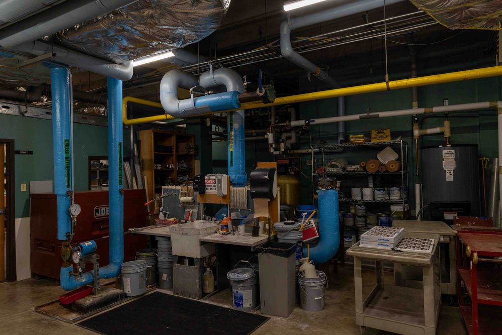 Industrial workshop interior with blue pipes, ventilation ducts, workbenches, and equipment in a dimly lit basement space.