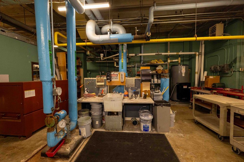 Industrial workshop with blue and yellow pipes, workbench with tools, buckets, and equipment in a utility room.
