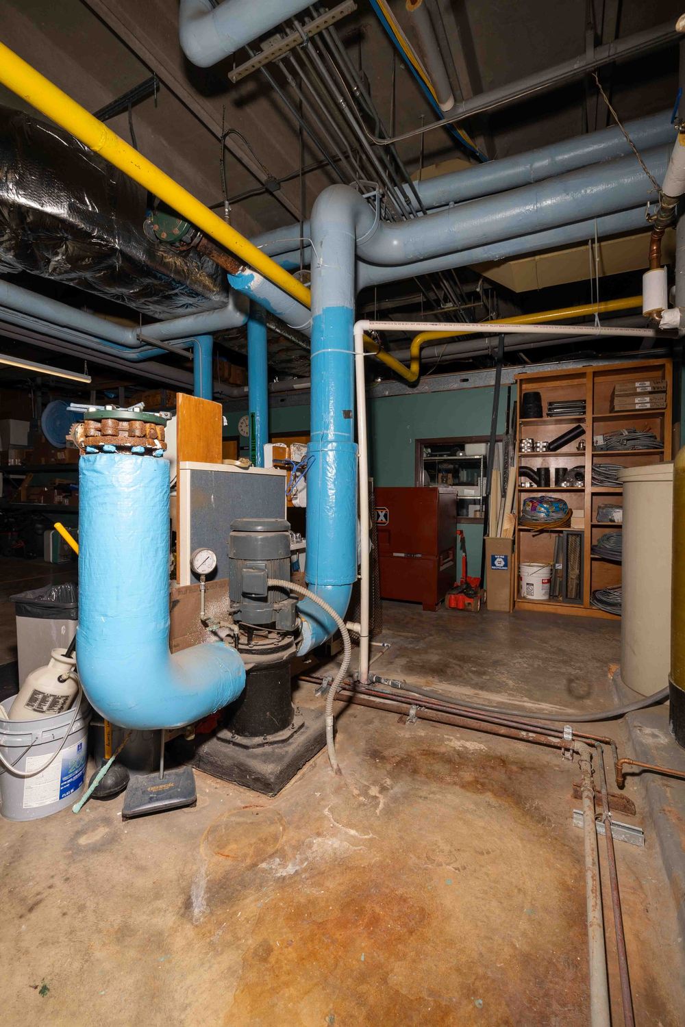 Industrial basement utility room with blue pipes, yellow gas lines, pump system, and wooden shelving unit on concrete floor.