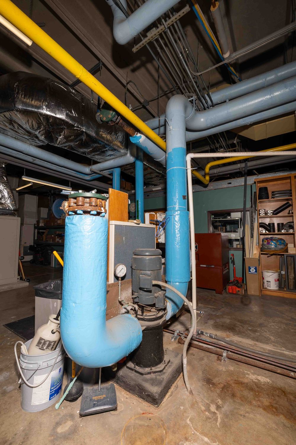 Industrial basement mechanical room with blue insulated pipes, yellow gas lines, pump equipment, and storage shelving.