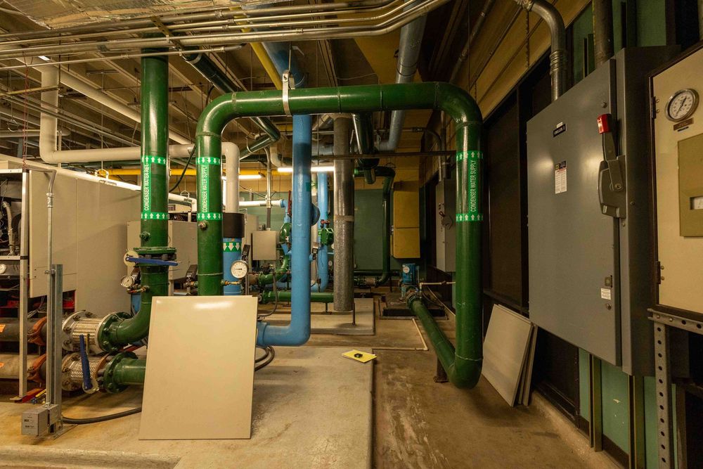 Green industrial pipes and mechanical equipment in a building utility room with electrical panels.