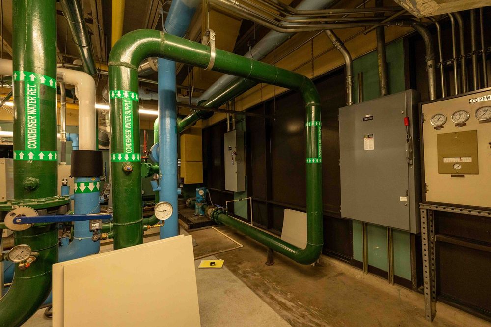Industrial mechanical room with green and blue pipes, HVAC systems, and electrical control panels in a commercial building.