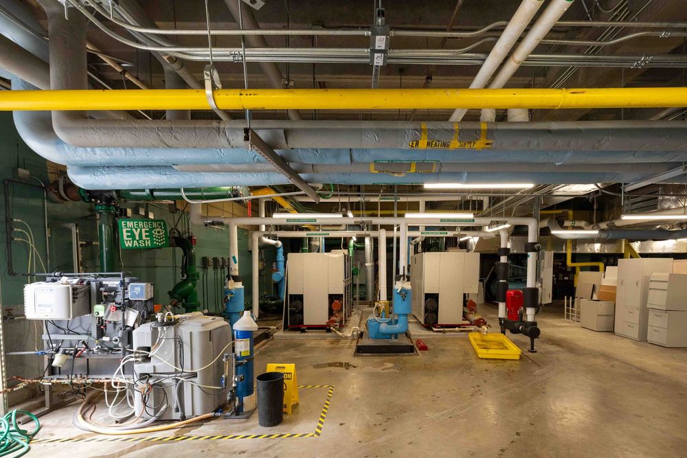 Large industrial mechanical room with yellow ceiling pipes, pumps, and equipment on concrete floor.