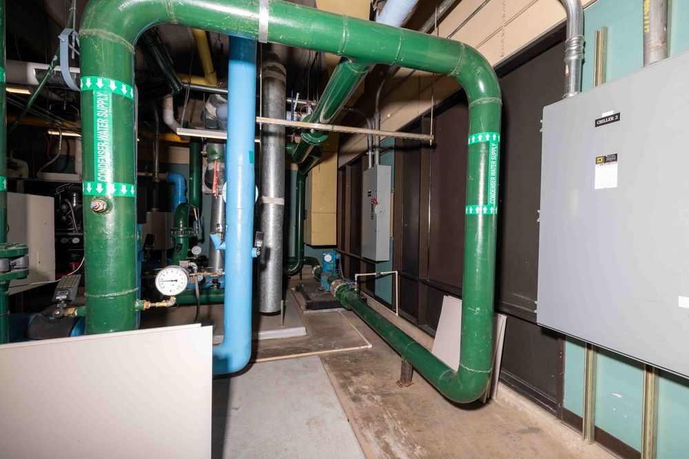 Industrial mechanical room with large green pipes, pumps, and electrical panels in a commercial building.