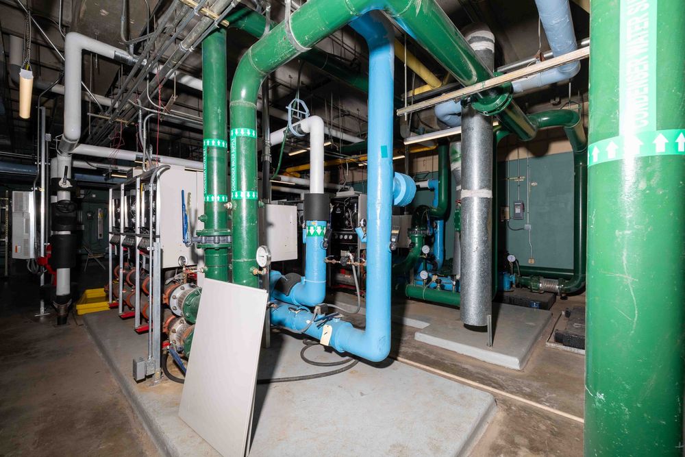 Industrial mechanical room with green and blue pipes, HVAC systems, and utility equipment in a commercial building.