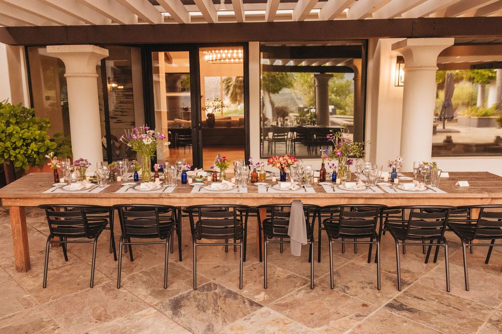 Elegant outdoor dining table set for a dinner party on a stone patio with floral centerpieces and black chairs.