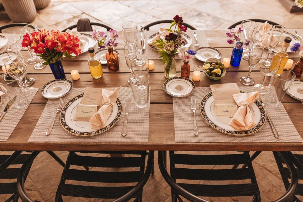 Elegant rustic wedding reception table set with floral centerpieces, fine china, crystal glasses, and linen napkins on a wooden farm table.