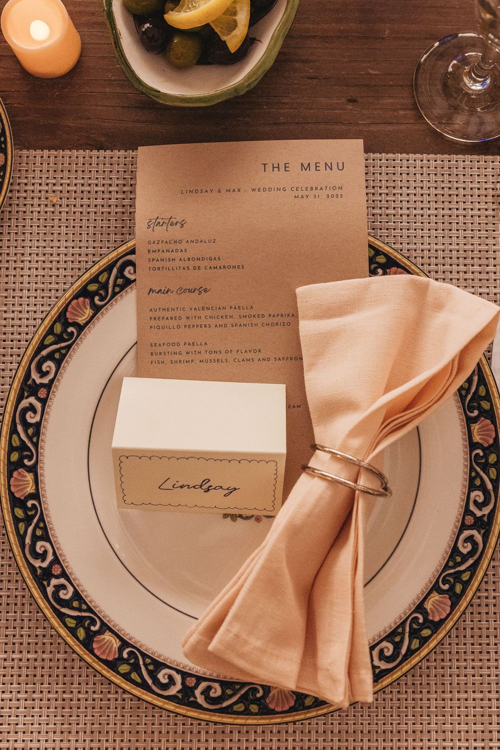 Elegant wedding place setting with a floral charger plate, peach napkin, gold ring, menu card, and name card reading Lindsay.