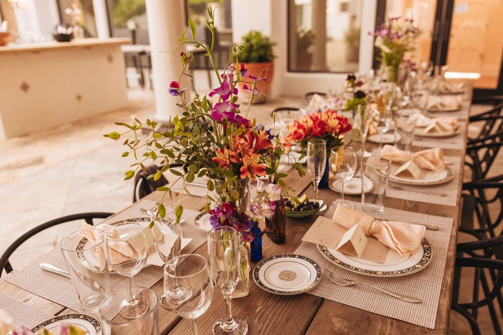 Elegant outdoor dining table set with colorful floral centerpieces, crystal glassware, and neatly folded napkins on decorative plates.