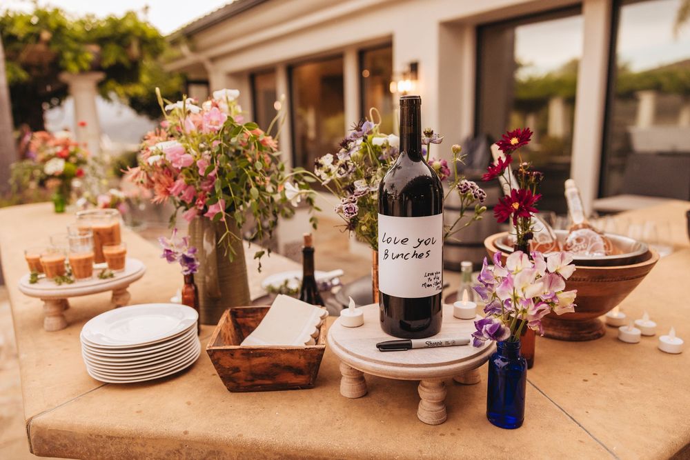 Outdoor wedding reception table with wine bottle, stacked plates, floral arrangements, and decorative wooden boxes.