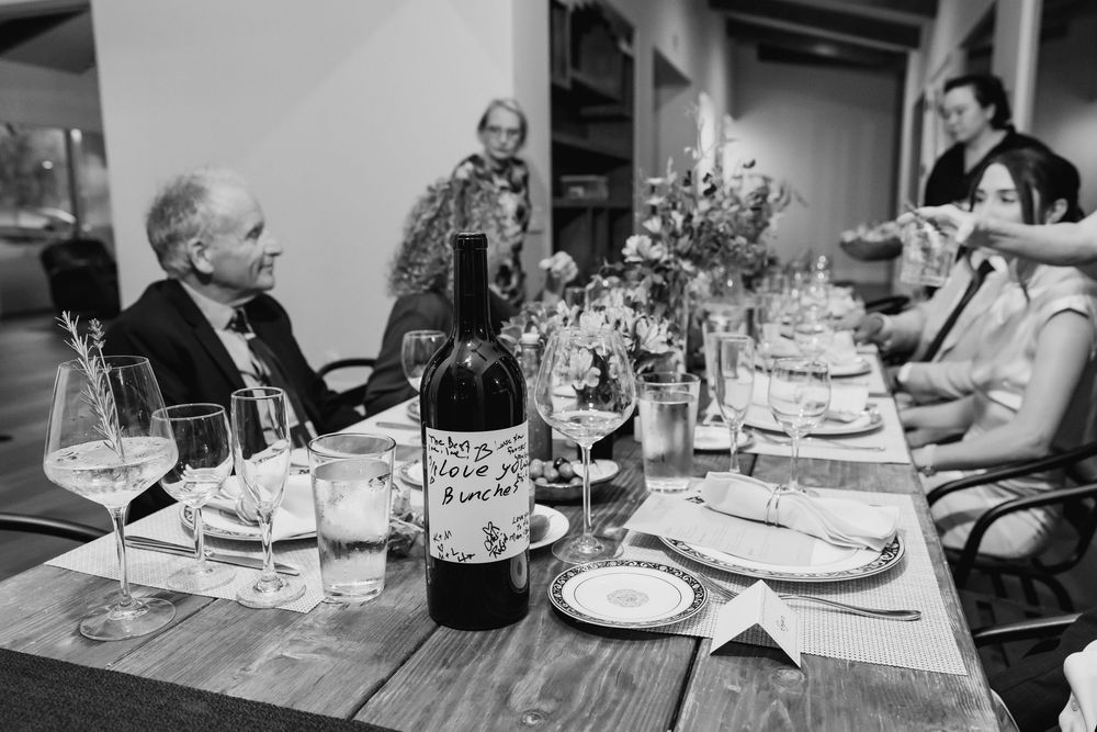 Black and white photo of guests dining at a rustic wooden table with wine glasses and a bottle of red wine.