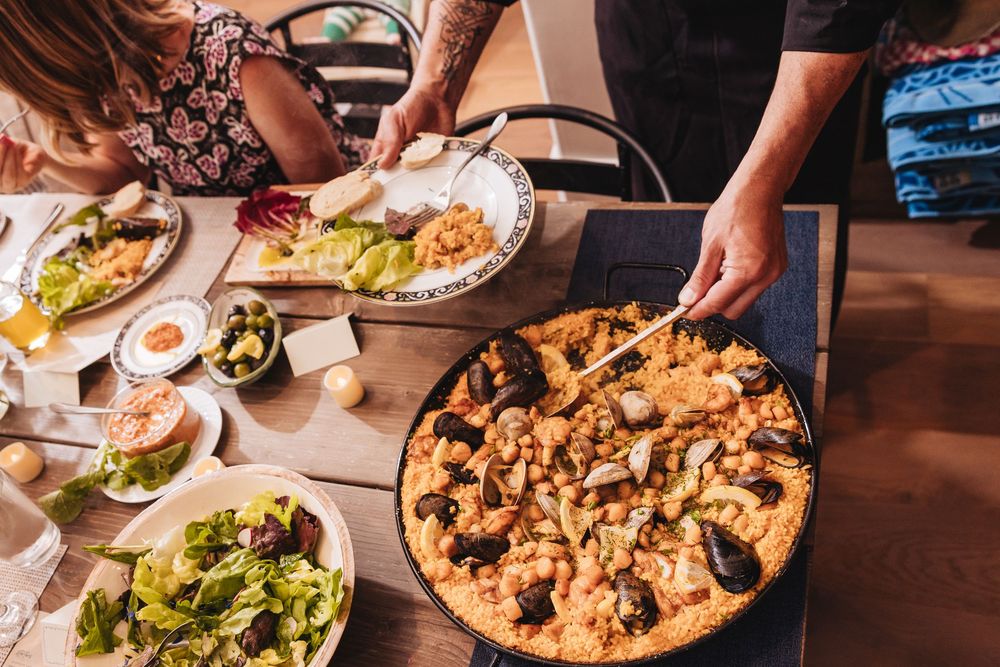People serving seafood paella with mussels and shrimp from a large pan at a rustic dining table.