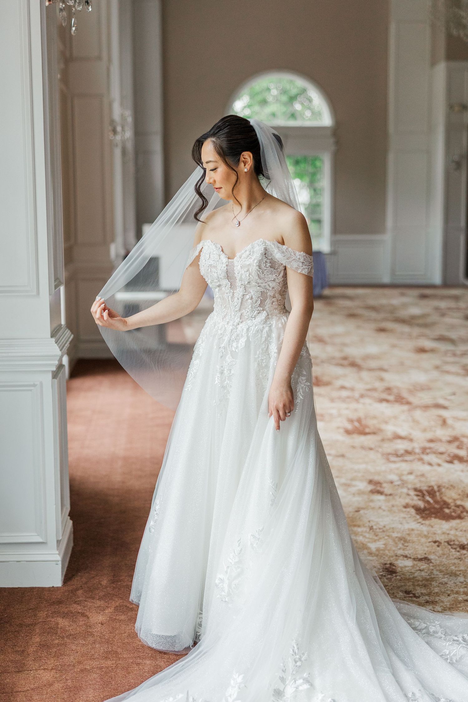 Bride in off-shoulder lace wedding gown with veil poses elegantly in a sunlit room with ornate decor.