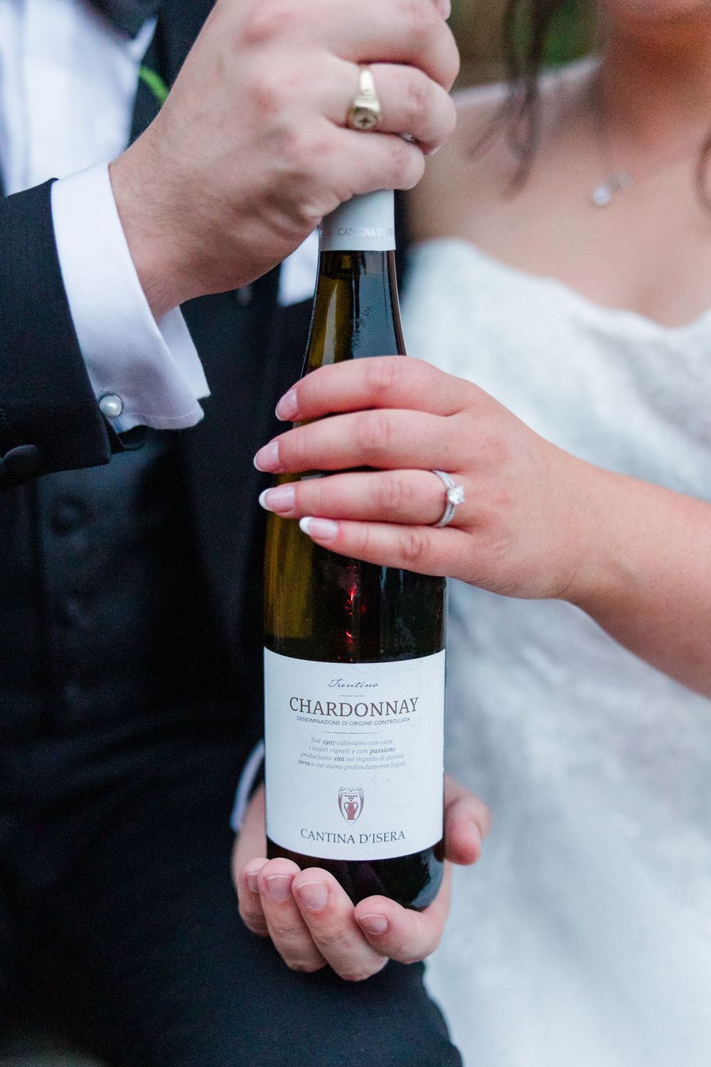 Newlyweds holding a Cantina D'Isera Chardonnay bottle, showcasing wedding rings on their hands.
