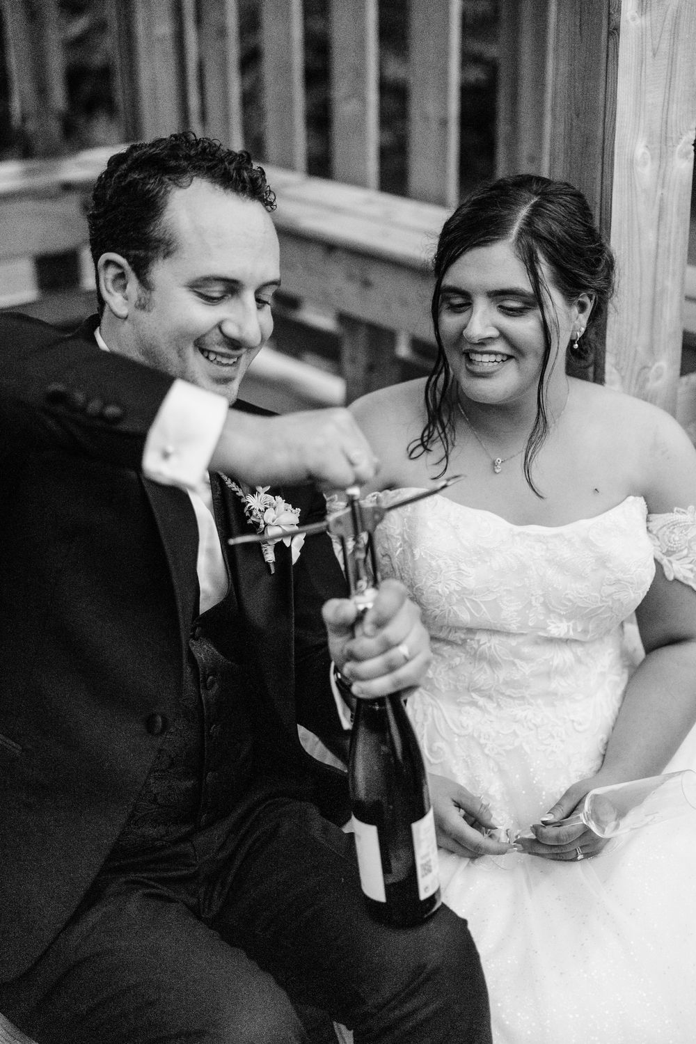 Black and white photo of a smiling bride and groom opening a champagne bottle together on their wedding day.