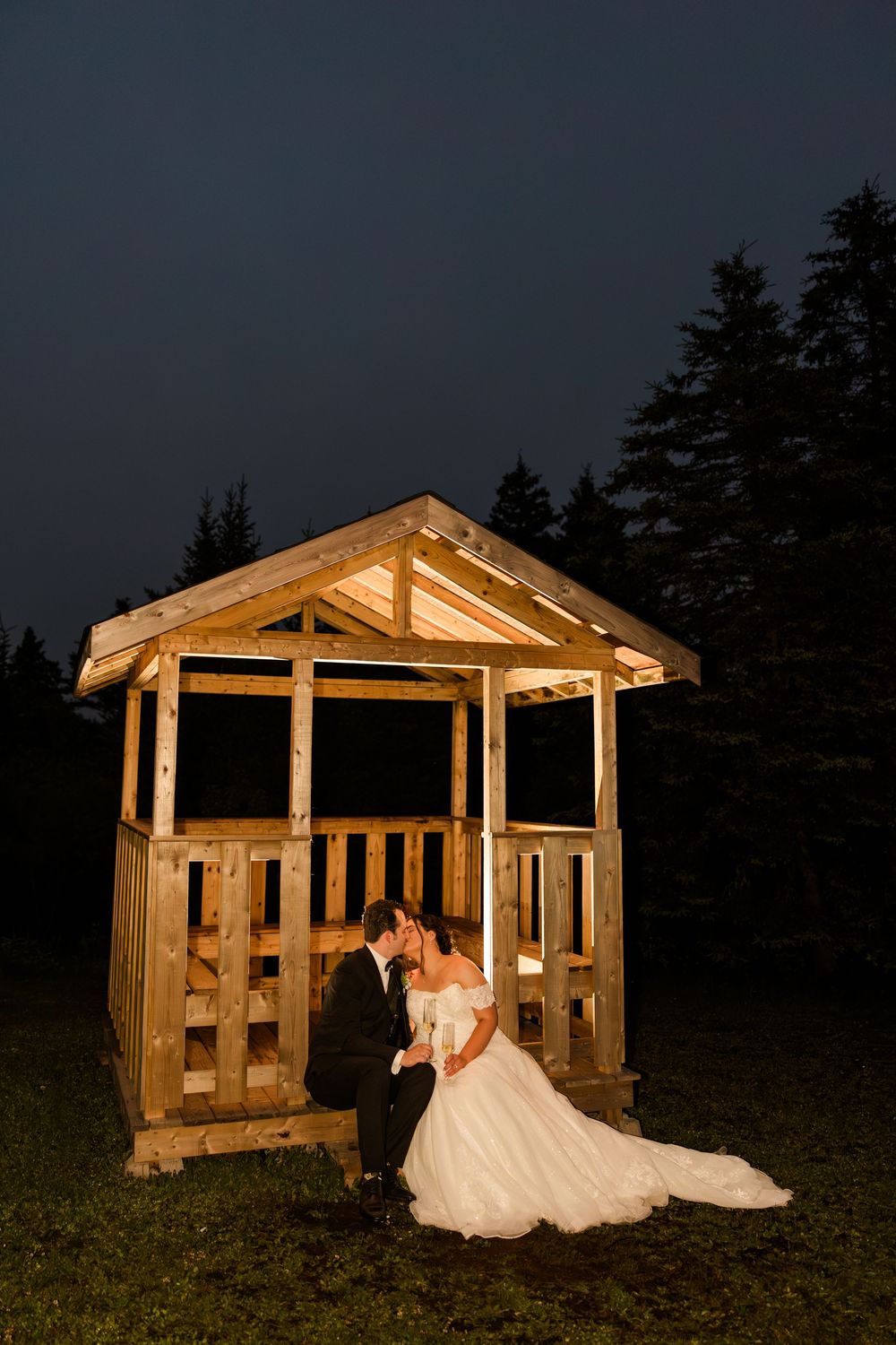 Bride and groom share a romantic kiss at night beside a rustic wooden gazebo surrounded by evergreen trees.