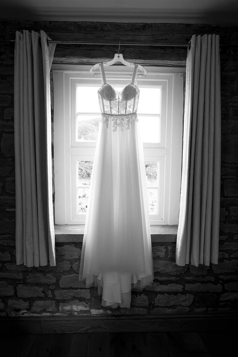 Black and white photo of an elegant wedding dress hanging on a hanger in front of a rustic stone wall window.